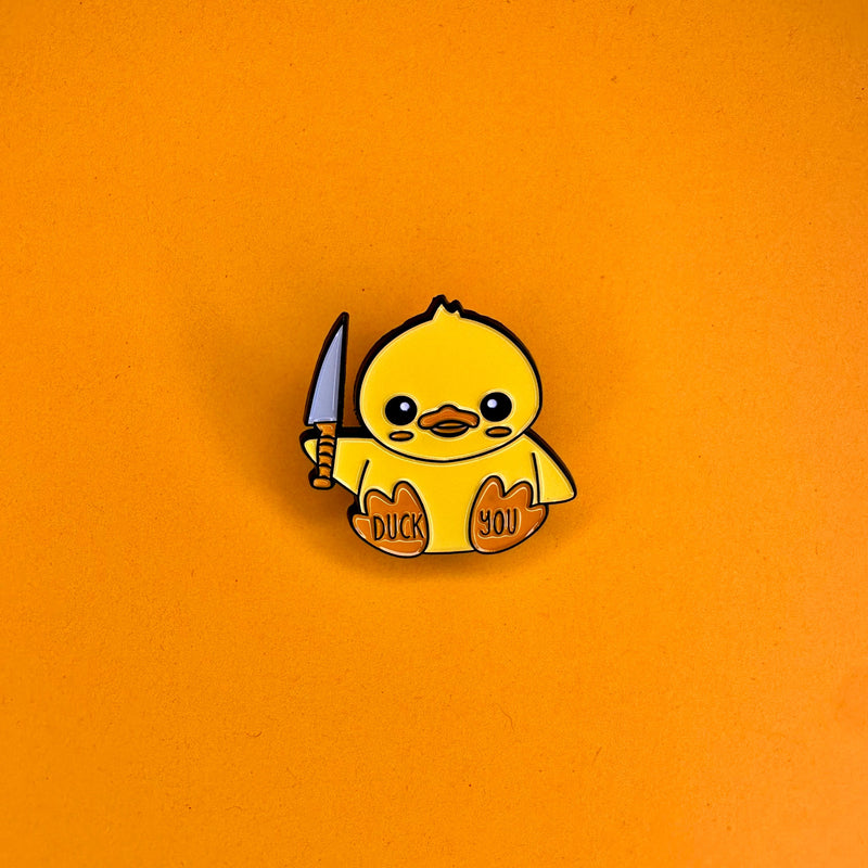 Enamel pin of a yellow duckling holding a silver knife, with the text 'Duck You' written across its orange chest, on an orange background - Pinp Me Up enamel pin