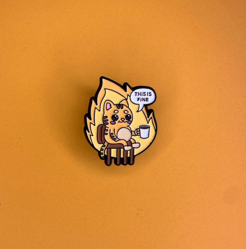 A small enamel pin shaped like a flame, featuring a cartoon cat sitting on a chair and holding a mug, with a speech bubble that says 'THIS IS FINE'. The pin is photographed on a solid orange background.